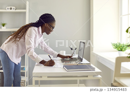 African American woman who works as accountant or secretary uses laptop and takes notes 134915483