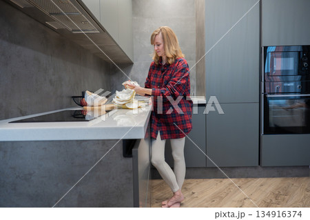 Blonde white woman preparing dinner in modern kitchen, slicing ingredients on counter and assembling meal. Casual plaid shirt, bare feet and relaxed posture convey comfortable domestic routine. Grey Blonde white woman preparing dinner in modern kitchen, slicing ingredients on counter and assembling meal. Casual plaid shirt, bare feet and relaxed posture convey comfortable domestic routine. Grey 134916374