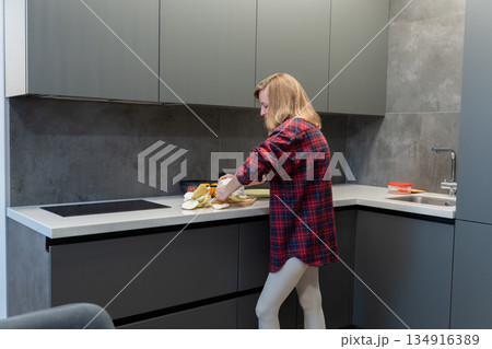 Woman arranging peeled pomelo on countertop in sleek kitchen, everyday domestic routine with relaxed posture and casual attire gray cabinets, bowl of fruit and soft ambient light emphasize home 134916389