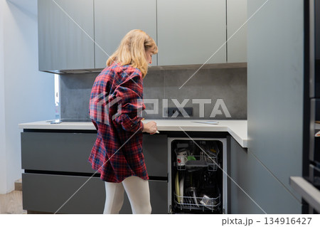Young white woman loading dishwasher in contemporary gray kitchen, wearing red plaid shirt, focused on placing glasses and plates into lower rack, minimalist cabinets and stainless countertop, natural Young white woman loading dishwasher in contemporary gray kitchen, wearing red plaid shirt, focused on placing glasses and plates into lower rack, minimalist cabinets and stainless countertop, natural 134916427