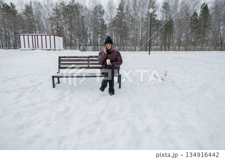 solitary woman sitting on snowy bench gazing into distance, wide winter landscape with sparse trees and modern building backdrop, mood of contemplation and quiet pause, footprints in snow and muted solitary woman sitting on snowy bench gazing into distance, wide winter landscape with sparse trees and modern building backdrop, mood of contemplation and quiet pause, footprints in snow and muted 134916442