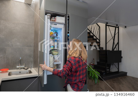 Woman opening refrigerator in modern kitchen, freelance designer grabbing quick snack between remote calls, wearing plaid shirt, blonde hair, openplan living area with staircase and media setup, Woman opening refrigerator in modern kitchen, freelance designer grabbing quick snack between remote calls, wearing plaid shirt, blonde hair, openplan living area with staircase and media setup, 134916460
