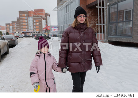 white mother and daughter walking on snowy apartment sidewalk, bundled in maroon puffer and pink jacket, holding hands, child carrying yellow shovel, city buildings and parked cars in background, white mother and daughter walking on snowy apartment sidewalk, bundled in maroon puffer and pink jacket, holding hands, child carrying yellow shovel, city buildings and parked cars in background, 134916461