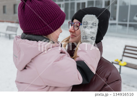 Closeup winter hug between grandmother and grandchild, mitten pressed to cheek, smiling adult wearing glasses and knit beanie, heavy puffer coats, soft snowfall, blurred urban background, intimate Closeup winter hug between grandmother and grandchild, mitten pressed to cheek, smiling adult wearing glasses and knit beanie, heavy puffer coats, soft snowfall, blurred urban background, intimate 134916510