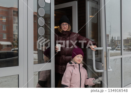 A woman and child exit a building, the mother leading the toddler through the glass entrance, dressed in winter coats and hats, smiling as they step out onto the sidewalk holding cupcakes 134916548