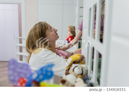 Caucasian mother helping daughter reach bookshelf in colorful nursery, organizing toys and choosing bedtime book, soft afternoon sunlight, warm smile, nurturing domestic scene, playful learning moment Caucasian mother helping daughter reach bookshelf in colorful nursery, organizing toys and choosing bedtime book, soft afternoon sunlight, warm smile, nurturing domestic scene, playful learning moment 134916551