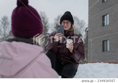 Woman kneeling filming child in snow, holding coffee and phone, child wearing purple hat playing with yellow shovel, apartment building and birch trees in background, candid urban winter family Woman kneeling filming child in snow, holding coffee and phone, child wearing purple hat playing with yellow shovel, apartment building and birch trees in background, candid urban winter family 134916587