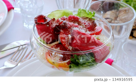Roasted beef slices nestling over crisp lettuce, garnished with cherry tomatoes and parmesan, presenting gourmet salad composition in clear glass serving bowl 134916595