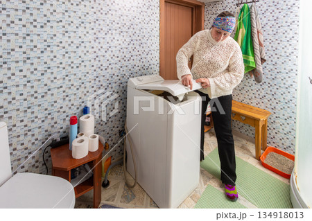 Washing clothes in vertical household washing machine, woman completes machine preparation, turning to leave area. Washing clothes in vertical household washing machine, woman completes machine preparation, turning to leave area. 134918013
