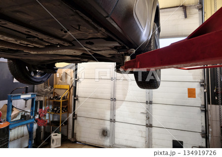 A car is raised by a hydraulic jack inside an auto repair shop. Tools and equipment are visible in the background. Light enters through the open door A car is raised by a hydraulic jack inside an auto repair shop. Tools and equipment are visible in the background. Light enters through the open door 134919726