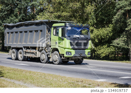 A green dump truck is driving on a road. Trees line the roadside. It is a sunny day in the countryside 134919737