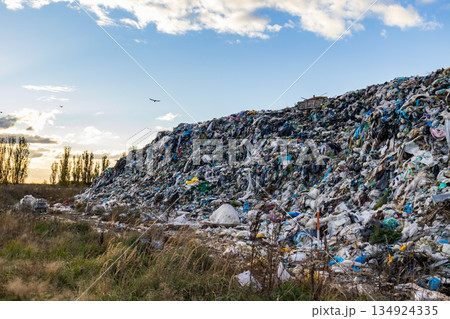 A large landfill contains heaps of garbage and waste materials under cloudy skies with some birds flying above the site 134924335