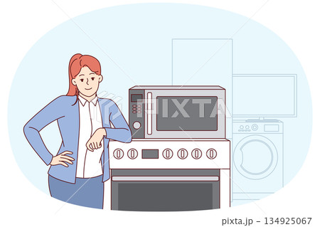Woman selling household appliances stands near oven and washing machine and looks at camera 134925067