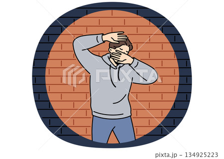 Man criminal in light of police lantern stands near brick wall and covers face with hands 134925223