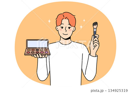 Non-binary man applies makeup and skin shadow, holding mirror and brush to apply powder 134925319