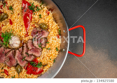 Top view of gourmet beef paella with roasted garlic and peppers in a traditional pan, with wide dark copy space on the right for text or design 134925580