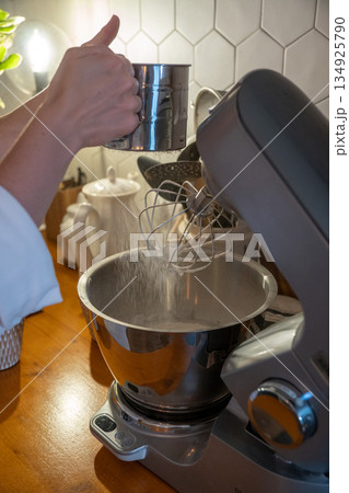 Hands sift flour into mixer bowl while preparing apple charlotte batter on wooden kitchen counter, capturing focused home baking moment 134925790