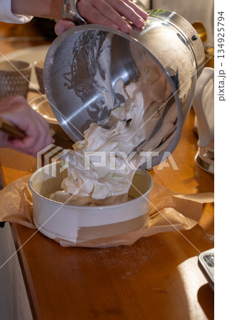 Hands pour airy apple charlotte batter from metal bowl into baking pan on kitchen counter, capturing cozy home baking moment with warm light Hands pour airy apple charlotte batter from metal bowl into baking pan on kitchen counter, capturing cozy home baking moment with warm light 134925794