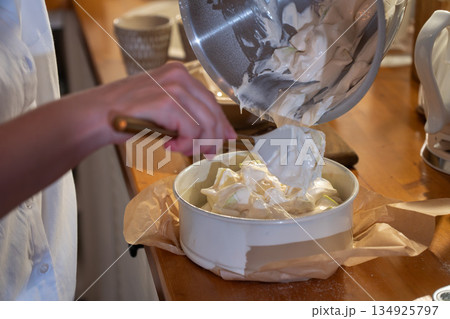Hands pour airy apple charlotte batter from metal bowl into baking pan on kitchen counter, capturing cozy home baking moment with warm light 134925797