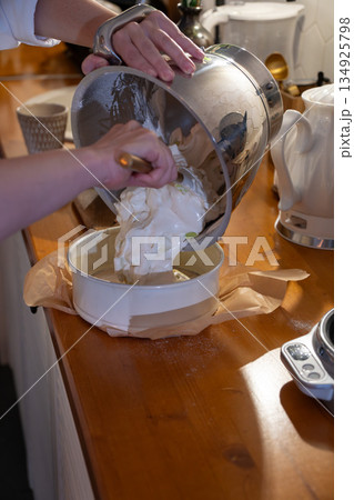 Hands pour airy apple charlotte batter from metal bowl into baking pan on kitchen counter, capturing cozy home baking moment with warm light Hands pour airy apple charlotte batter from metal bowl into baking pan on kitchen counter, capturing cozy home baking moment with warm light 134925798