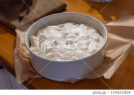 Ready apple charlotte batter rests in round baking pan lined with parchment on wooden kitchen counter, showing smooth creamy texture with apple pieces 134925809
