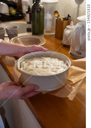 Hands hold baking pan filled with creamy apple charlotte batter on a kitchen countertop, showing quiet home baking moment in warm indoor light Hands hold baking pan filled with creamy apple charlotte batter on a kitchen countertop, showing quiet home baking moment in warm indoor light 134925810