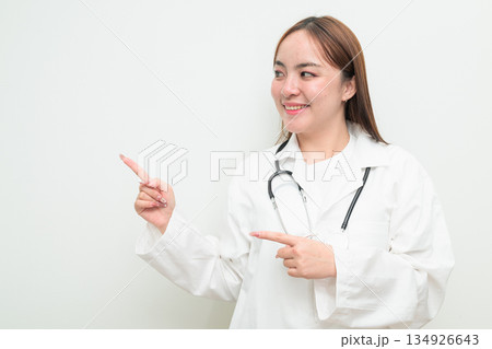 Portrait of young Asian female doctor against white background pointing finger Portrait of young Asian female doctor against white background pointing finger 134926643