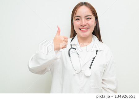 Portrait of young Asian female doctor against white background giving thumbs up gesture Portrait of young Asian female doctor against white background giving thumbs up gesture 134926653