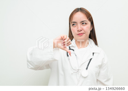Portrait of young Asian female doctor against white background giving thumbs down gesture Portrait of young Asian female doctor against white background giving thumbs down gesture 134926660