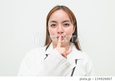 Portrait of young Asian female doctor against white background finger on lips Portrait of young Asian female doctor against white background finger on lips 134926667