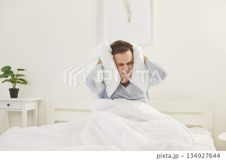 Sleepy angry upset young man sitting on bed covering head with pillows suffering from loud sound. 134927644