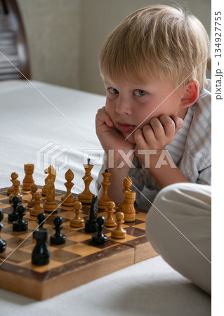 Young boy rests his chin on his hands while studying chessboard at home, showing focus, curiosity, thoughtful mood in soft natural light Young boy rests his chin on his hands while studying chessboard at home, showing focus, curiosity, thoughtful mood in soft natural light 134927755