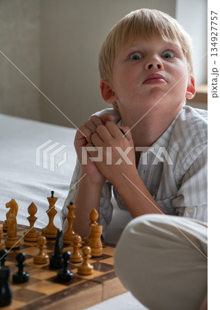 Young boy reacts emotionally while playing chess at home, showing surprise, curiosity with expressive eyes in soft natural daylight 134927757