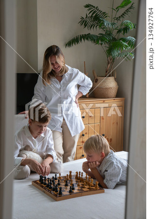 Mother stands beside her children playing chess on a bed, creating a warm family moment with soft daylight, neutral tones, and a calm home atmosphere Mother stands beside her children playing chess on a bed, creating a warm family moment with soft daylight, neutral tones, and a calm home atmosphere 134927764