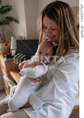Mother gently holds her baby at home, smiling while the infant looks at the camera, creating a warm, intimate family moment in soft natural light Mother gently holds her baby at home, smiling while the infant looks at the camera, creating a warm, intimate family moment in soft natural light 134927767
