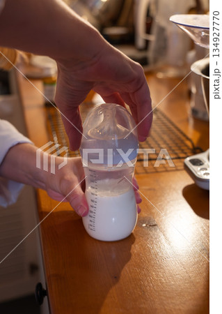 Hands prepare a baby bottle with milk on a wooden kitchen counter, showing careful pouring and a calm everyday parenting routine in warm light Hands prepare a baby bottle with milk on a wooden kitchen counter, showing careful pouring and a calm everyday parenting routine in warm light 134927770