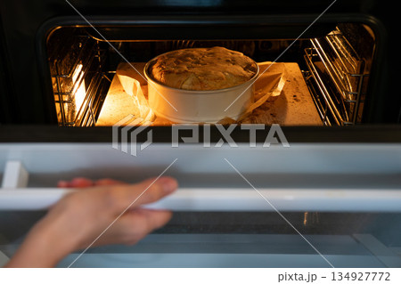 Apple charlotte cake bakes in an oven inside a round pan, lit by warm oven light, showing rising dough and a cozy homemade baking mood Apple charlotte cake bakes in an oven inside a round pan, lit by warm oven light, showing rising dough and a cozy homemade baking mood 134927772