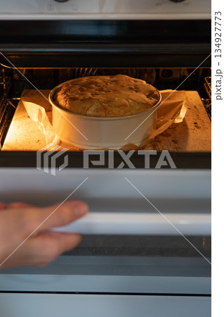 Apple charlotte cake bakes in an oven inside a round pan, lit by warm oven light, showing rising dough and a cozy homemade baking mood 134927773