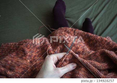 Melange brown sweater with knitting needles and women legs in velvet socks on dark green background. Knitting needles and balls of warm melange yarn. Girl legs and knitting work in process.  134928257