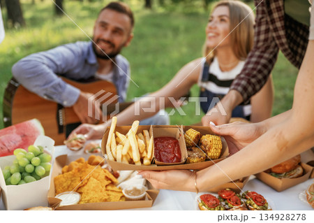 Sitting by the table. Group of friends are having picnic on the field with food in eco boxes Sitting by the table. Group of friends are having picnic on the field with food in eco boxes 134928278