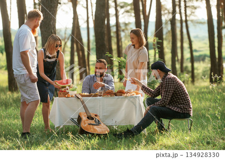 Trees at background. Group of friends are having picnic on the field with food in eco boxes Trees at background. Group of friends are having picnic on the field with food in eco boxes 134928330