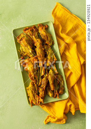 Delicious deep-fried zucchini flowers are neatly arranged on a green platter, showcasing their crispy texture. A bright yellow napkin adds warmth to the scene Delicious deep-fried zucchini flowers are neatly arranged on a green platter, showcasing their crispy texture. A bright yellow napkin adds warmth to the scene 134928921