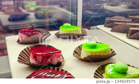 Assorted mini mousse cakes with bright pink and green mirror glaze topped with small macarons on display in a bakery 134929575