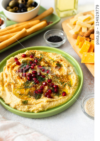 Delight in a classic hummus dish topped with pomegranate seeds, served alongside a variety of cheeses, nuts, and breadsticks perfect for sharing during gatherings Delight in a classic hummus dish topped with pomegranate seeds, served alongside a variety of cheeses, nuts, and breadsticks perfect for sharing during gatherings 134929779