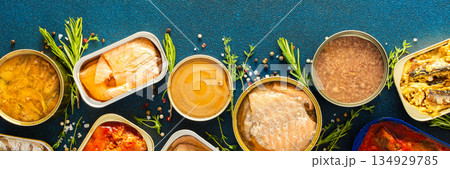 A top view of open tin cans with canned salmon, mackerel, sprat, sardine, tuna, herring, and fish pate, surrounded by spices and herbs on a blue background, banner 134929785