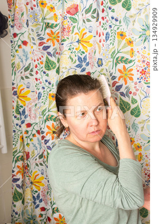 In a bright room decorated with a colorful floral curtain, a 50-year-old woman calmly brushes her hair as part of her morning routine, showcasing a peaceful start to her day 134929909