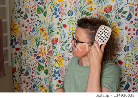 A 50-year-old woman holds a hairbrush while looking in the mirror during her morning bathroom routine. She checks her hair near a floral shower curtain A 50-year-old woman holds a hairbrush while looking in the mirror during her morning bathroom routine. She checks her hair near a floral shower curtain 134930039