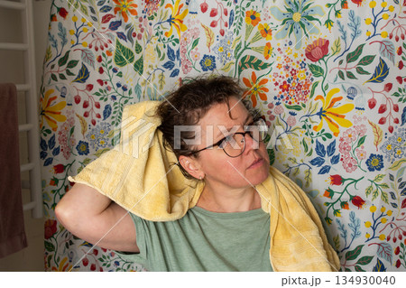 A 50-year-old woman starts her morning routine in the bathroom. She looks at herself in the mirror while holding a towel over her shoulder. The colorful background adds brightness 134930040