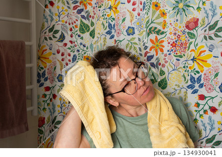 A 50-year-old woman starts her morning routine in the bathroom. She looks at herself in the mirror while holding a towel over her shoulder. The colorful background adds brightness A 50-year-old woman starts her morning routine in the bathroom. She looks at herself in the mirror while holding a towel over her shoulder. The colorful background adds brightness 134930041