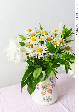 Fresh daisies are arranged in a decorative vase, brightening up a light room. The soft pink tablecloth enhances the cheerful atmosphere, showcasing spring flowers 134930207